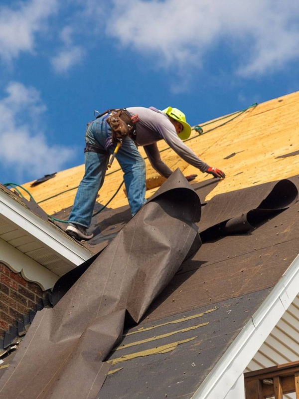 A construction worker in a white hard hat, blue jacket, safety harness, and gloves kneels on a shiny metal roof, using a power drill to secure a panel under a clear blue sky with scattered clouds and a distant horizon.