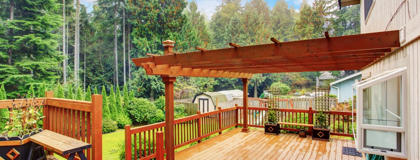 Wooden deck with pergola, potted plants, and railing overlooking a green backyard with trees and a shed.