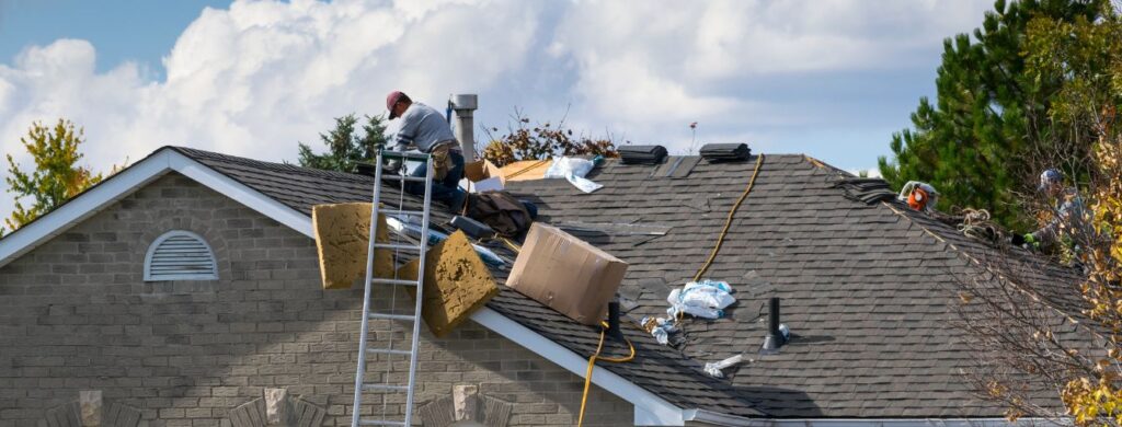 Two workers repairing a damaged asphalt shingle roof on a brick house, one in a red cap climbing a ladder with yellow insulation board